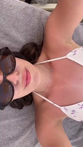 The video shows a close-up overhead shot of a woman lying on a gray towel, possibly on a beach or by a pool. She is wearing sunglasses and a white bikini with a floral pattern. Her skin appears tanned and slightly reddish, suggesting she may have been in the sun. The woman is looking up at the camera and smiling faintly. A thin silver necklace with a small pendant is visible around her neck. The lighting suggests it is daytime.
