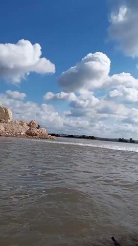 The video shows a body of water with ripples and small waves. On the left side of the frame, there is a rocky shore with some larger rocks and debris. In the distance, the water extends to a horizon with a treeline and hills. The sky is blue with fluffy white clouds. The perspective is low, as if the camera is at water level or just above it. The overall scene depicts a natural outdoor environment, possibly a lake or a wide river, under a partly cloudy sky.