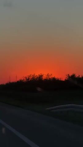 The video captures a vibrant sunset scene from a moving vehicle. The sky displays a striking array of colors, with intense orange and red hues dominating the horizon, gradually fading into a muted grey-blue at the top. Silhouetted against the bright sky are a line of trees or bushes that run across the middle of the frame. To the left, several power line pylons are also visible in silhouette. The bottom portion of the frame shows a dark grey road with a white solid line on its left edge, and a red and white road marker post is present on the right side of the road. A guardrail can be seen along the roadside on the left. The scene evokes the peaceful end of a day. No visible text is present.
