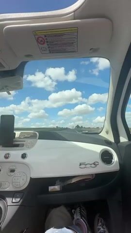 The video shows the interior of a white Fiat 500. The camera is positioned in the passenger seat, looking out the windshield at a bright blue sky with scattered white clouds. A road stretches into the distance, with fields on either side. In the foreground, the dashboard and steering wheel are visible. A person's legs and feet, wearing sneakers, are visible in the lower part of the frame. The camera then pans to the right, showing the car door and a side mirror. Finally, the camera turns to capture a young woman in the passenger seat, wearing a pink top and sunglasses. She is sticking out her tongue and holding up two fingers in a peace sign. Her hair is blowing in the wind. The sun is shining brightly, and the sky remains visible throughout the clip.