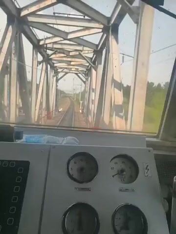 The video shows a view from the inside of a train as it travels along a track. The train is passing over a steel girder bridge. The landscape is green with trees lining the tracks. The train's controls are visible. There are several dials and gauges as well as a keypad display showing a speed of 60 km/h. There is an alarm sound.