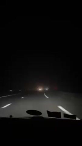 The video shows a nighttime view from inside a car driving on a highway. The road is black and marked with white lane lines. There are other vehicles ahead, visible as blurred headlights and red taillights in the distance. A windshield sticker is visible in the bottom center of the frame. The overall lighting is poor, and the conditions appear foggy or hazy. The audio includes music.