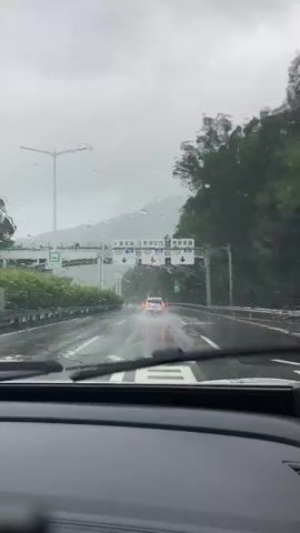 The video is a first-person perspective shot from inside a car driving on a wet highway in rainy weather. The windshield is speckled with raindrops, some of which distort the view. Ahead, a white car is visible driving in the same direction, its taillights reflecting on the wet road. Overhead, there are highway signs and streetlights. To the sides of the highway, there are guardrails and lush green trees on a hillside. The sky is overcast and gray, indicating continuous rain. The road is wet and reflects the car's headlights. The overall atmosphere is gloomy due to the weather conditions.