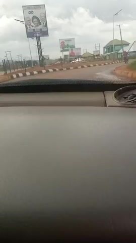 The video is taken from inside a vehicle, looking out through the windshield. The dashboard is visible in the foreground. Outside, there is a road with buildings and billboards on either side. There is a motorcycle in the distance. The weather appears to be cloudy. The video captures a short clip of the vehicle's movement.