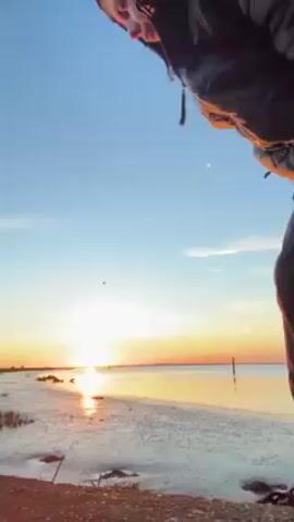 A person in a dark puffer jacket with the hood up, black pants, and black shoes stands by a body of water at sunset. The sky is blue fading into orange near the horizon. The water reflects the orange sunlight. The person is facing the water, their back to the camera. The tide appears to be low, exposing mudflats.