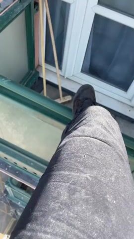 The video appears to be shot from a high balcony, looking down and then up along the side of a multi-story building. Initially, the camera focuses on a person's leg and foot, standing on the edge of the balcony. The view then shifts to showcase the construction of the balcony itself, showing stacked layers with a green metal frame and glass panels. Looking down, cars are visible parked on the street below. As the camera pans up, the structure of the building is revealed. There are multiple balconies with a rough, reddish-brown finish.  A set of ropes with an "ASAP LOCK" device is rigged along the side of the building, extending to the top, which is marked with bird spikes. The sky above is clear and blue.