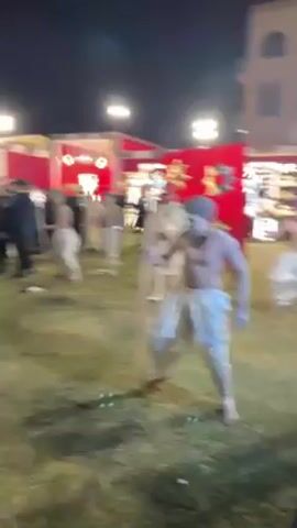 The video shows a group of men in silver body paint and traditional Indian clothing dancing on a lawn at night. They are mostly shirtless, wear matching pants and have their faces painted white with dark markings. Several of them carry wooden staffs, while others carry decorative red banners with gold embellishments. Behind the dancers, there is a crowd of onlookers, including people in formal attire. The setting appears to be an outdoor event, possibly a wedding or festival, with decorative structures and lighting visible in the background. The lawn is well-lit and manicured.