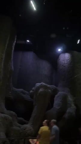 The video shows a low-angle, shaky shot of a dark indoor environment that appears to be a themed attraction or exhibit. Large, textured structures resembling rocks or ancient pillars dominate the foreground and mid-ground, with some parts illuminated by spotlights. In the background, more structures are visible, along with what looks like scaffolding or support beams. Several people are present, but they are out of focus and their faces are not clearly visible. The overall atmosphere is dim and somewhat mysterious, with artificial lighting creating dramatic shadows. The camera movement is erratic, suggesting it's being held by someone walking or moving through the space.