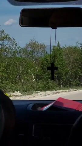 The video captures a scenic daytime drive from the perspective of a vehicle's front passenger seat or dashboard. Through the windshield, a rearview mirror hangs prominently, from which a black cross charm dangles. Partially visible on the dashboard below are a red book or folder and other indistinct items.

The car appears to be navigating a winding paved road through a lush, green, mountainous or hilly landscape. Dense trees and bushes, many with bright green spring foliage and some with yellow blossoms, line both sides of the road. In the mid-ground, a tall cellular or communication tower is visible among the trees. The background features rolling hills and distant mountains under a clear blue sky with scattered white clouds. White road markers or short guardrail posts are visible at the edge of the road as it curves. The bright sunlight suggests good weather conditions.

The scene depicts a peaceful journey through a rural, verdant area, consistent with locations like Grab, Bosnia and Herzegovina. No specific individuals or readable text are visible in the frame.