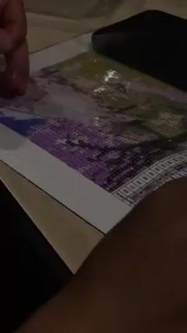 A person is working on a diamond painting craft project. The project depicts a purple and green image. A black smartphone is resting on top of the craft. The person uses a stylus tool to apply the small diamond-like beads to the adhesive canvas, following the symbols and color key provided along the edge of the canvas. The video is shot in low light.