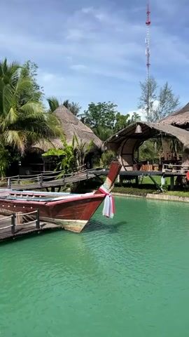 A tranquil scene unfolds at a waterfront resort in Tambon Pa Kae Bo Hin, Thailand. The turquoise water of a calm lagoon is dotted with a fountain, its spray catching the sunlight. Moored to a wooden dock is a longtail boat, its vibrant red hull adorned with a colorful fabric lei.

Behind the boat, a series of charming thatched-roof bungalows and open-air structures are nestled amongst lush greenery. These buildings, built on stilts, feature natural materials like bamboo and wood, contributing to the tropical ambiance. Bridges connect some of the structures, allowing easy access over the water.

A tall telecommunications tower stands in the background, a modern contrast to the traditional architecture. The sky above is a clear blue, with a few wispy clouds, suggesting a pleasant, sunny day. The overall atmosphere is one of peaceful relaxation and natural beauty, characteristic of a tropical getaway. No people are visible in the frame.