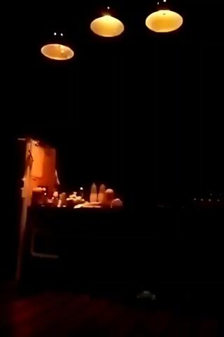 The video shows a dark room with three pendant lights hanging from the ceiling. Beneath the lights, there is a table displaying various items, possibly food or decorative objects. The overall scene is dimly lit, with only the lights and the objects on the table visible. The table appears to be near a wall. In the background, there are some distant lights.