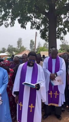 The video shows two clergymen dressed in white robes with purple stoles, standing outside beneath a tree while conducting what appears to be a funeral service. The clergyman on the left is holding a book and has his right hand raised. Both clergymen are looking at the book, but the clergyman on the left is speaking. The crowd behind the clergymen is blurry, but you can see that there are several people standing around the clergymen. The environment is sunny and appears to be an outdoor area with some houses in the distance.