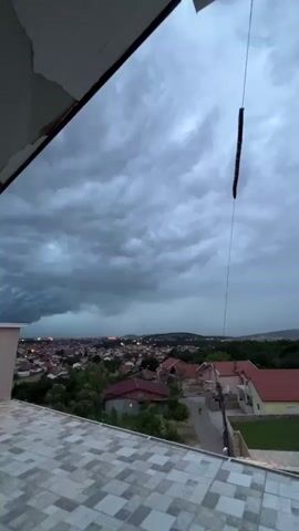 The video, likely filmed from a rooftop in Podgorica, Montenegro, captures a dramatic stormy sky over a town at dusk. Dark, heavy storm clouds dominate the sky, particularly on the left side where they appear most ominous, suggesting an approaching or ongoing downpour. Below, the sprawling urban landscape of Podgorica is visible, with numerous houses featuring red-tiled roofs and streetlights beginning to illuminate the evening.

In the immediate foreground, a tiled rooftop patio extends from a building. To the right, a thin string or wire hangs from above, with a long, dark, segmented object resembling a centipede or similar insect suspended from it, swaying gently. The distinct sound of strong wind is audible throughout the clip, adding to the atmosphere of an impending storm. Further down in the town, a single person can be seen walking on a street. The overall impression is one of anticipation as the storm front moves across the landscape.