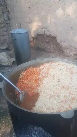 A large pot of food is being cooked outdoors over an open fire. The pot is filled with rice on the right side and carrots and other ingredients on the left side. A metal ladle is in the pot, stirring the food. There is a large metal container in the background and a wall made of mud or plaster behind that. The ground is dirt and stones. The fire is visible at the bottom of the pot.