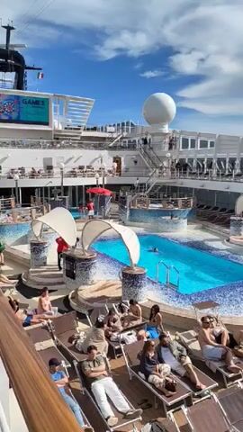 The video shows an outdoor pool deck on a cruise ship. Multiple people are lounging on deck chairs, some sunbathing and others conversing. A bright blue pool with a mosaic tile design is in the center, with a person swimming in it. Overhead, there's a deck above with additional seating. The sky is blue with some clouds, and various ship structures are visible in the background, including a large white dome-shaped structure. The overall atmosphere is relaxed and leisure-oriented.