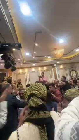 The video shows a group of men inside a banquet hall, possibly at a wedding or celebration. Most of the men are dressed in traditional clothing, including headscarves. Several men are clapping, and others are gathered in a small cluster. A man in a pink shirt appears to be taking pictures or videos. Professional lighting and camera equipment are visible on the ceiling. The overall atmosphere appears lively and celebratory.