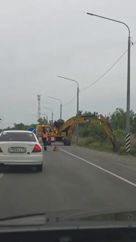 From the perspective of a car approaching a road construction zone, a construction worker in an orange vest and yellow hardhat stands on the road. He is in front of a barrier with a detour sign, a “Road Work” sign, and traffic cones. An idle Caterpillar excavator is behind him, partially blocking the road. A white car is stationary nearby, on the same side of the road. The sky is overcast, and there are tall light poles and a cell tower visible in the background. Trees and bushes are present on the right side of the road.