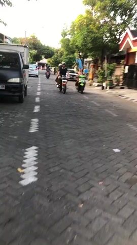 The video shows a street scene with several motorcycles. In the foreground, the pavement is made of dark gray interlocking bricks arranged in a herringbone pattern. White markings are painted on the road. There are buildings and trees on both sides of the street. 

In the center of the frame, two people are riding on a motorcycle. The rider in front is wearing a dark t-shirt, shorts, and a helmet. The passenger behind them is wearing a light-colored t-shirt and shorts. They are moving away from the camera.

To the right of this motorcycle, another motorcycle is visible, ridden by a single person wearing a light-colored t-shirt and shorts. This motorcycle is green.

Further down the street, two more motorcycles are visible, also moving away from the camera. One is being ridden by a single person, and the other by two people.

On the left side of the street, a white pickup truck is parked. Buildings line the right side of the street, one of which has an orange and white facade. The lighting suggests it is either early morning or late afternoon. The overall impression is a casual street scene in a populated area, possibly in Southeast Asia given the presence of multiple motorcycles.
