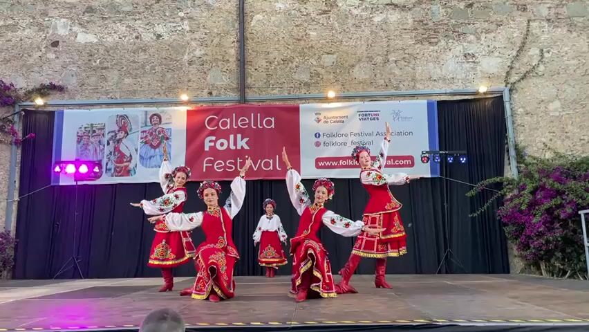 A group of five female dancers in traditional red and white embroidered costumes perform on a wooden stage in front of a banner that reads "Calella Folk Festival." The dancers have their hair tied back and are wearing floral headbands. Their costumes consist of white blouses with intricate embroidery on the sleeves and chest, red vests, and red skirts with yellow and black trim. They are also wearing tall red boots. The dancers are positioned in a semi-circle, with one dancer in the center and two on each side. They are all in motion, with their arms raised and legs bent. The background is a stone wall, and there are purple flowers growing on the right side of the stage. The lighting on the stage is provided by spotlights.