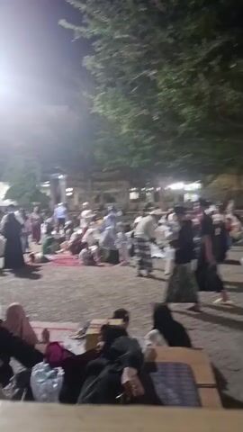 The video shows a nighttime scene with many people gathered outdoors. The ground appears to be paved. There are trees and buildings in the background. Many individuals are sitting or lying on the ground, some on mats or blankets. There are also some people standing and walking. The lighting suggests artificial light sources such as lamps or streetlights. The individuals appear to be dressed in traditional clothing, with many women wearing headscarves. Some people are carrying bags or other belongings. The atmosphere seems to be one of waiting or gathering.