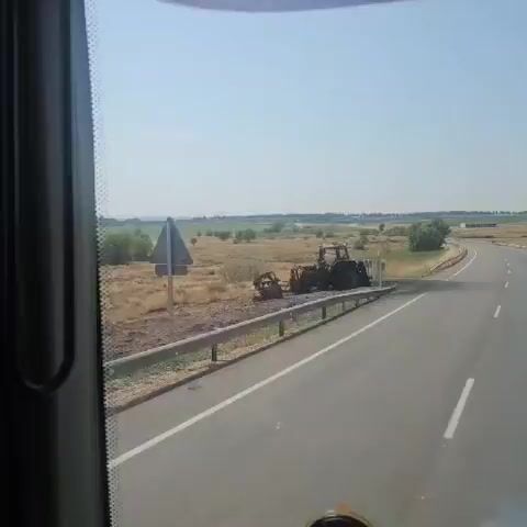 The video shows a landscape with a road and a damaged tractor. The tractor is dark in color and appears to be burnt out, with debris around it. It is located on the side of the road, next to a metal guardrail. Beyond the road and the tractor, there is dry, grassy terrain with scattered trees and bushes. In the distance, there is a flat landscape that might be a field or a plain, stretching towards the horizon under a clear, bright sky. The camera is positioned from inside a vehicle, suggesting the video was recorded while moving. There are no people visible in the video.