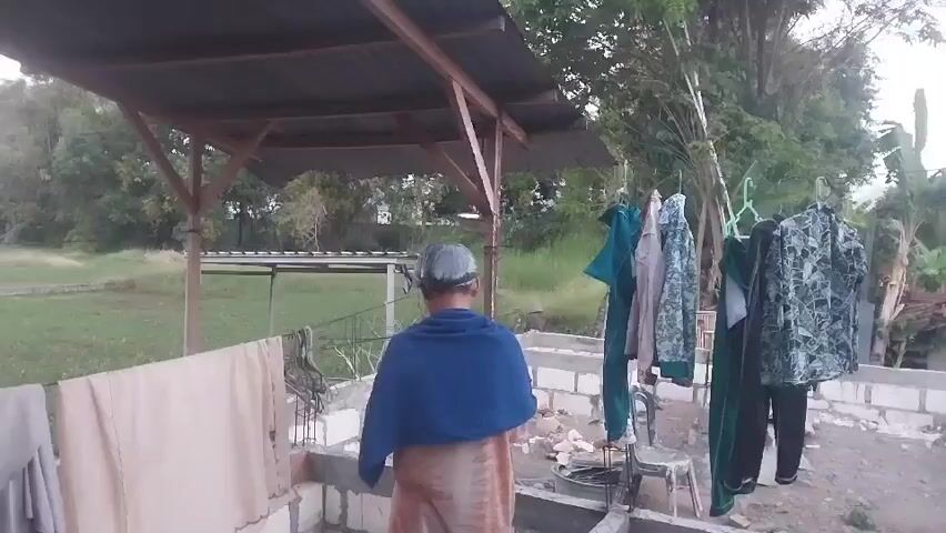 A person wearing a blue cloth over their shoulders and a patterned skirt is standing in front of a stone wall. They are facing away from the camera and appear to be washing or wringing out a piece of clothing. To the left of the person, there is a clothesline with light-colored fabric hanging on it. To the right, several pieces of clothing are hanging on hangers on a clothesline, including green and patterned garments. Behind the person and the clothesline is a covered structure with a corrugated metal roof, supported by wooden beams. The environment appears to be outdoors, with trees and grassy areas visible in the background. The ground is uneven and appears to be a mix of dirt and rubble.