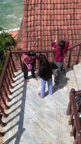 The video captures a high-angle view of three young individuals on an outdoor staircase. The staircase has a marbled surface and is bordered by a wooden railing with decorative posts. The individuals are positioned on the stairs, facing each other. The person on the left, wearing a maroon shirt and black pants, holds a bouquet of purple flowers. The person in the center, wearing a black top and blue jeans, has long dark hair and stands with their back to the camera. The person on the right, in a maroon long-sleeved shirt and grey pants, appears to be dancing or gesturing with their hands. Behind them, a red tiled roof dominates the background, and beyond that, a choppy, turquoise ocean is visible under a bright sky. The lighting suggests it is daytime, with distinct shadows cast on the stairs.