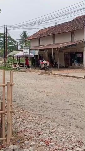 A person wearing a red jacket and black pants is seen pushing a motorcycle with a basket on the front. The motorcycle is positioned on a dirt road in front of a building with a white awning and a sign that reads "GALUNGGANG BAKSO". The building appears to be a shop or restaurant. To the left of the building, there is a covered seating area with a pink and white umbrella. The road is unpaved and appears to be under construction, with loose gravel and debris. In the foreground, there is a bamboo fence. The sky is overcast.