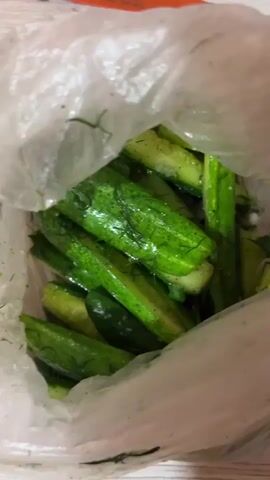 A hand is shown interacting with a clear plastic bag filled with chopped cucumbers and dill. The bag is placed on a wooden surface next to a knife with an orange blade and a white handle. The cucumbers are green and appear to be seasoned. The hand moves the bag, causing the contents to shift.