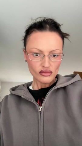 The video features a medium shot of a woman indoors. She has dark hair pulled up in a messy bun, light skin, and is wearing glasses with gold rectangular frames. Her makeup includes defined brows, blush, and dark lipstick. She wears small flower-shaped earrings, a black t-shirt, and a zipped-up gray hoodie. The background shows a room with a white ceiling and light-colored walls. There is a light fixture on the ceiling, and a window is visible with greenery outside. She adjusts her glasses with her right hand. The camera angle is slightly low, looking up at the subject.