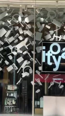 The video shows the front of a store called “Daily.”  The store front consists of a large glass window decorated with outlines of common objects, such as scissors, glasses, cell phones, and flashlights. At the center of the window is a black square with the word "Daily" in white text.  Inside the store, shelves of various items can be seen. The camera is stationary and zooms out slightly.