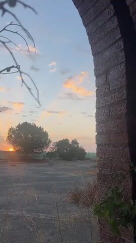 A video captured at sunset shows the sun setting behind a tree, casting rays of light through the clouds. The scene is framed by an arched brick structure on the right and branches of a tree in the foreground on the left. The ground appears to be a mix of asphalt and dry grass.