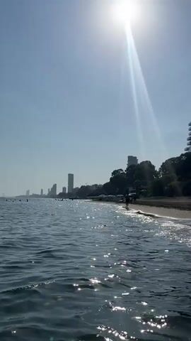 The video shows a view from the water looking towards a beach and buildings. The sun is high in the sky and creates a glare on the water. There are several tall buildings visible along the coastline, some of which appear to be hotels or apartment complexes. The beach is sandy and there are a few people visible, some walking and others sitting on the beach. The water is calm and there are small waves breaking on the shore.