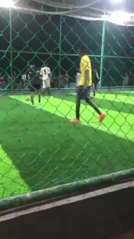 A video shows a fenced-in indoor soccer field with green artificial turf. Several people are playing or watching. The players visible wear assorted clothing, including black shorts and white jerseys, and a yellow jersey with blue trim. One player is dribbling a soccer ball. The field is lit by overhead lights. A portion of the fence is visible in the foreground, partially obstructing the view. The camera is presumably held by a spectator positioned outside the fence.