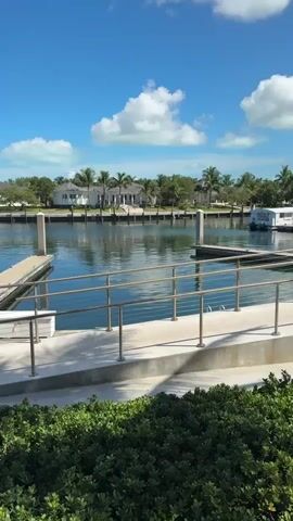 The video shows a tranquil harbor on a sunny day. The sky is a brilliant blue with scattered white clouds. Calm, clear water reflects the sky and the surrounding scenery. Several boats are docked or cruising in the distance, including a sailboat on the left and a motor yacht in the center. A wooden dock extends into the water, with metal railings along its edges. Along the shoreline, there are modern houses surrounded by palm trees and lush green vegetation. The scene conveys a sense of peace and luxury.