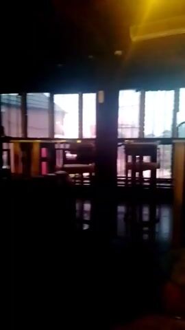 The video shows a dimly lit room, possibly a restaurant or bar. There are large windows visible with light coming through, and some tables and chairs. As the camera pans, a light fixture and a blurry view of a bar with shelves of bottles can be seen. The overall atmosphere is dark and somewhat blurry.