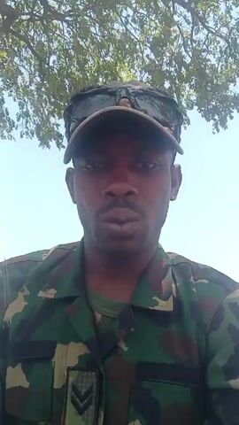 A man in a military uniform, camouflage pattern, and a baseball cap with sunglasses on top, is looking at the camera. He has a mustache and a small, vertical scar under his lower lip. He is under a tree with sparse green leaves, against a clear, bright sky.  The man moves his mouth into a pucker repeatedly.