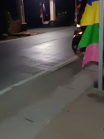 A motorcycle with a rider is seen at night on a paved road. The motorcycle's headlights are on, illuminating the road. The rider is wearing a headscarf and patterned clothing. The motorcycle is black with red accents on the front. The road has a white line marking the edge. There are colorful flags on the right side of the frame. In the background, there are buildings and streetlights. The scene appears to be in an urban or suburban area.