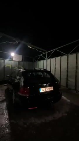 The video shows a car wash area with a black Volkswagen Golf station wagon. The car's license plate reads "A 8324 HP." The video then transitions to a gas station at night. A man and a woman are standing outside the gas station. The gas station is brightly lit with fluorescent lights under the canopy. A black car is parked at one of the pumps.