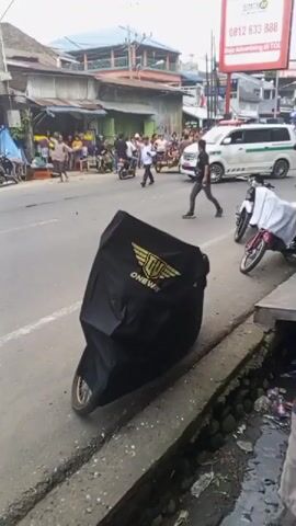 A motorcycle covered in a black tarp with a gold logo is parked on the side of a road. People are standing around it and on the sidewalk. A white van with green accents is driving in the background. A group of people are gathered on the sidewalk in the distance, and a few are standing in the street. The road appears to be cracked and uneven. There is a small drainage ditch to the right of the motorcycle.
