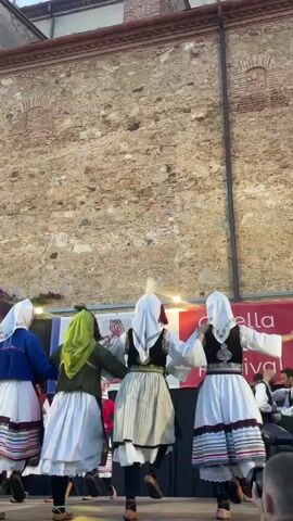 Four dancers in traditional folk costumes are performing on a stage in front of a rustic stone building. The dancers are all women, dressed in white skirts with colorful trim, embroidered vests, and headscarves. They are holding hands and moving in a circular formation, their backs mostly to the camera. The costumes appear to be vibrant and detailed, suggesting a cultural performance.

Behind the dancers, a large banner is visible with text that reads "Calella Folk Festival." This indicates the event taking place is a folk festival in Calella, Spain. To the right of the dancers, several men in similar traditional attire are standing, possibly musicians or other performers.

The scene is outdoors, and the lighting suggests it is daytime, possibly late afternoon or early evening, as there are some artificial lights illuminating the stage area. The stone building behind the stage has an aged appearance with visible brickwork and architectural details. The overall atmosphere seems festive and celebratory, consistent with a cultural festival.