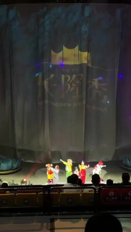 A stage show is in progress. Six costumed performers stand under spotlight, on a stage with a gray backdrop displaying "CHIMELONG SHOW HENGQIN ZHUHAI" in Chinese and English. The audience sits in the foreground, partially visible. The performers wear brightly colored outfits, reminiscent of circus or clown attire. Stage lighting includes green and blue accents. A sign indicating "no smoking" in Chinese is attached to the seating.