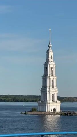 A bell tower stands on a small island in a large body of water. The tower is white and has a cross on top. The water is blue and there are trees on the shore in the distance. The sky is clear and blue. A large, golden, disc-shaped object is visible in the distance, rising above the trees.