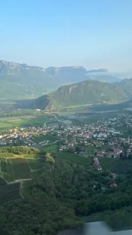 This is a vertical, slightly shaky video showing a scenic view of a valley with mountains in the background. In the foreground, there are steep hillsides covered with rows of vineyards. A small town or village with red-tiled roofs is visible in the mid-ground, nestled within the valley. Beyond the town, there are agricultural fields, appearing as patchwork squares of green. The mountains in the distance are large and imposing, with some cloud cover around their peaks. The sky is a clear, pale blue, suggesting it is daytime, likely early morning or late afternoon due to the soft lighting. The perspective suggests the footage is being captured from a moving vehicle, possibly a train or car, as evidenced by the slight motion blur and the presence of a railing or edge in the very bottom of the frame. There are no people or animals visible in the frame. The overall impression is one of serene rural landscape.