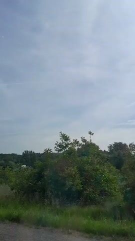 The video captures a drive through a rural landscape with a bright, overcast sky. The scene is dominated by green fields and trees. In the distance, a few houses are visible, some with red roofs, nestled amongst the foliage. One house appears to have solar panels on its roof, suggesting a sustainable approach to energy. A white, greenhouse-like structure is also present in one of the fields.

The main object of interest is a large billboard advertising McDonald's. It features an arrow pointing upwards and the text "9 km MOP Widoma," indicating a McDonald's restaurant is 9 kilometers away at a rest stop. Below this, another billboard is partially visible, with Polish text that reads "JUŻ OTWARTE," meaning "OPEN NOW."

The overall impression is a journey through a peaceful, rural area, with the McDonald's advertisement serving as a modern commercial marker within the natural setting. The weather appears to be mild, with a lot of cloud cover, suggesting it's daytime, likely morning or afternoon. There are no people visible in the frame, and the activity is simply the movement of the vehicle capturing the footage. The location is confirmed as Michałowice, Poland, which aligns with the Polish text on the billboard.