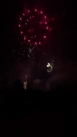 The video captures a fireworks display at night. Silhouetted figures, presumably a crowd, are visible in the foreground. Several fireworks explode in the dark sky, illuminating the scene with bursts of green, white, and red light. Some fireworks create a circular pattern, while others are more scattered. Smoke from the explosions drifts through the air. The overall atmosphere is festive and dark.