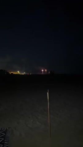 The video shows a nighttime view of a beach with fireworks in the distance. The foreground is dark, showing a sandy beach with a blanket and a wooden pole. In the background, there is a dark horizon line with some lights that appear to be from buildings or a town. The main focus of the video is a series of fireworks exploding in the sky, primarily in shades of yellow and green. The fireworks are spaced out, with two large yellow bursts near the center and smaller bursts of green and yellow scattered around them. The sky is dark, indicating that it is nighttime.