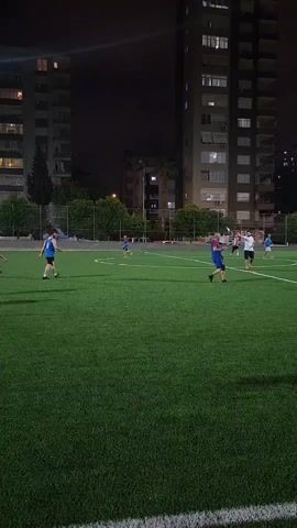 The video shows an outdoor soccer field at night. There are several people on the field, some of whom appear to be playing soccer. The field is lit by artificial lights, and there are buildings in the background. One person in the foreground is wearing a white shirt and black shorts and appears to be stretching or warming up. Another person further back is wearing a blue shirt and black shorts and is facing away from the camera. There are other people scattered across the field, also in athletic wear. The overall scene suggests a casual game or practice session taking place in an urban environment after dark.