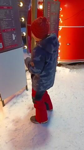A young boy stands in the snow, facing a menu board and glancing at it. He is wearing a red knit hat, a camouflage jacket, red snow pants, and grey boots. The menu board is black and white, listing food and prices in Russian. Behind the menu board is a red food truck with strings of lights. The ground is covered in snow. There's music playing in the background.